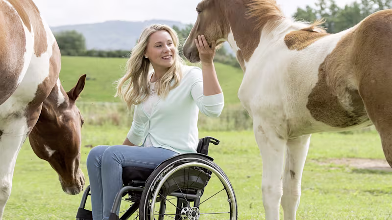 A woman in an Ottobock wheelchair is smiling in a meadow surrounded by two horses and pets one of them on the neck