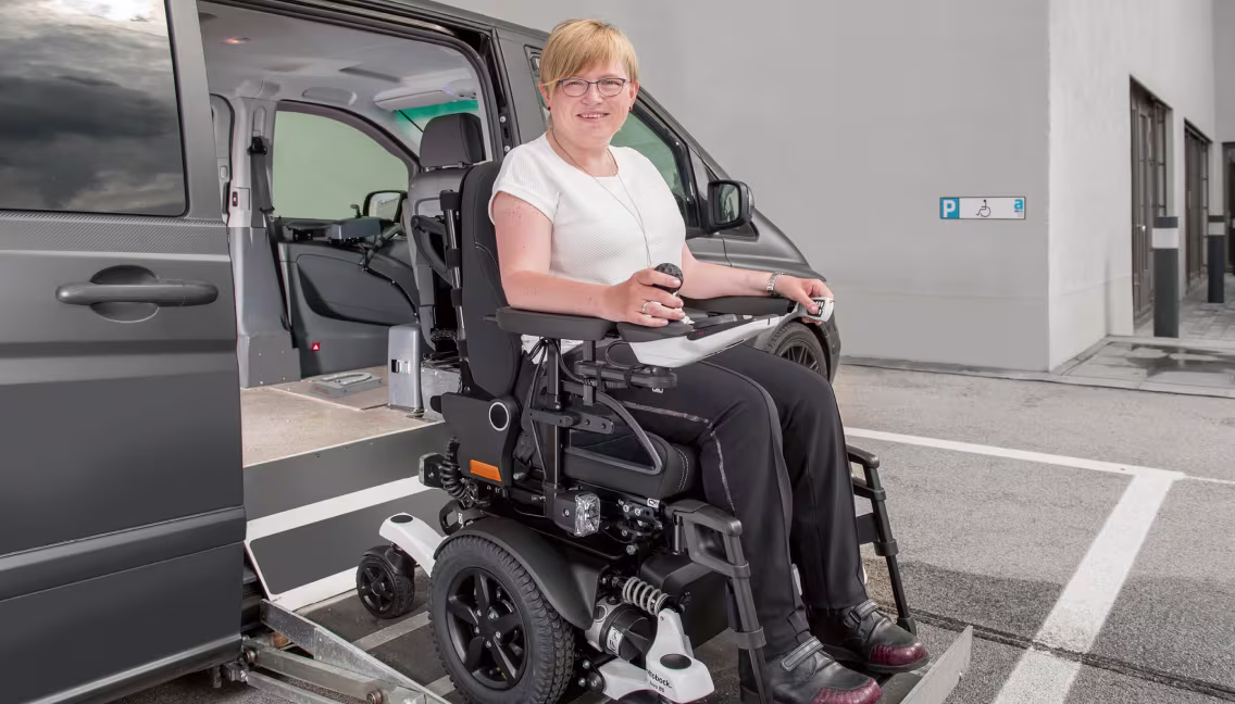 A middle-aged woman in a Juvo B5/B6 wheelchair uses a mechanical lift to enter the side door of a van.