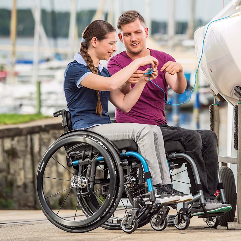 A woman and a man in Ottobock wheelchairs at a harbor in front of a small boat that is lifted up
