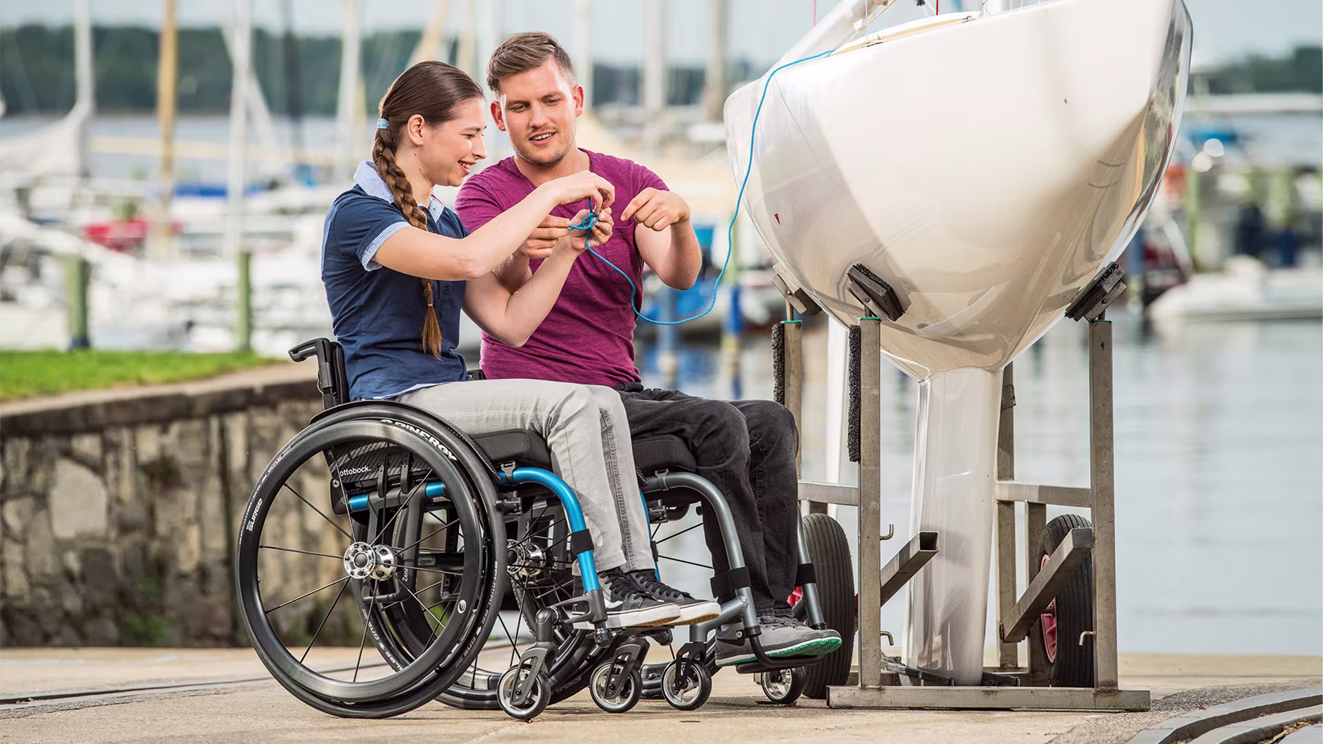 A woman and a man in Ottobock wheelchairs at a harbor in front of a small boat that is lifted up