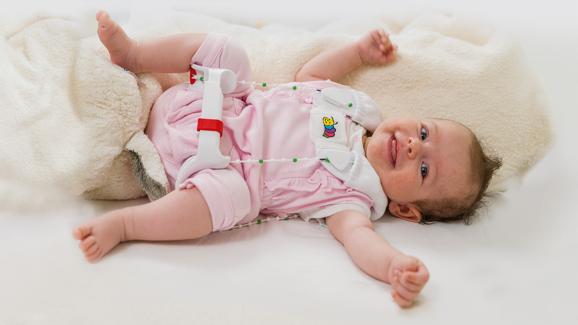 An infant in pink pajamas lays on a fuzzy blanket wearing the Ottobock Tubingen hip flexion and abduction orthosis 