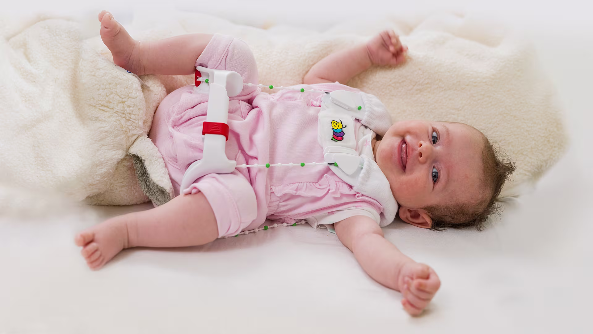 An infant in pink pajamas lays on a fuzzy blanket wearing the Ottobock Tubingen hip flexion and abduction orthosis