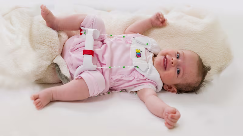 An infant in pink pajamas lays on a fuzzy blanket wearing the Ottobock Tubingen hip flexion and abduction orthosis