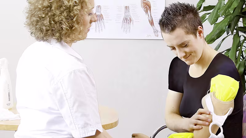 Eine Ottobock Mitarbeiterin sitzt vor einer Frau, die mit ihrer rechten Hand ihre linke Armprothese berührt