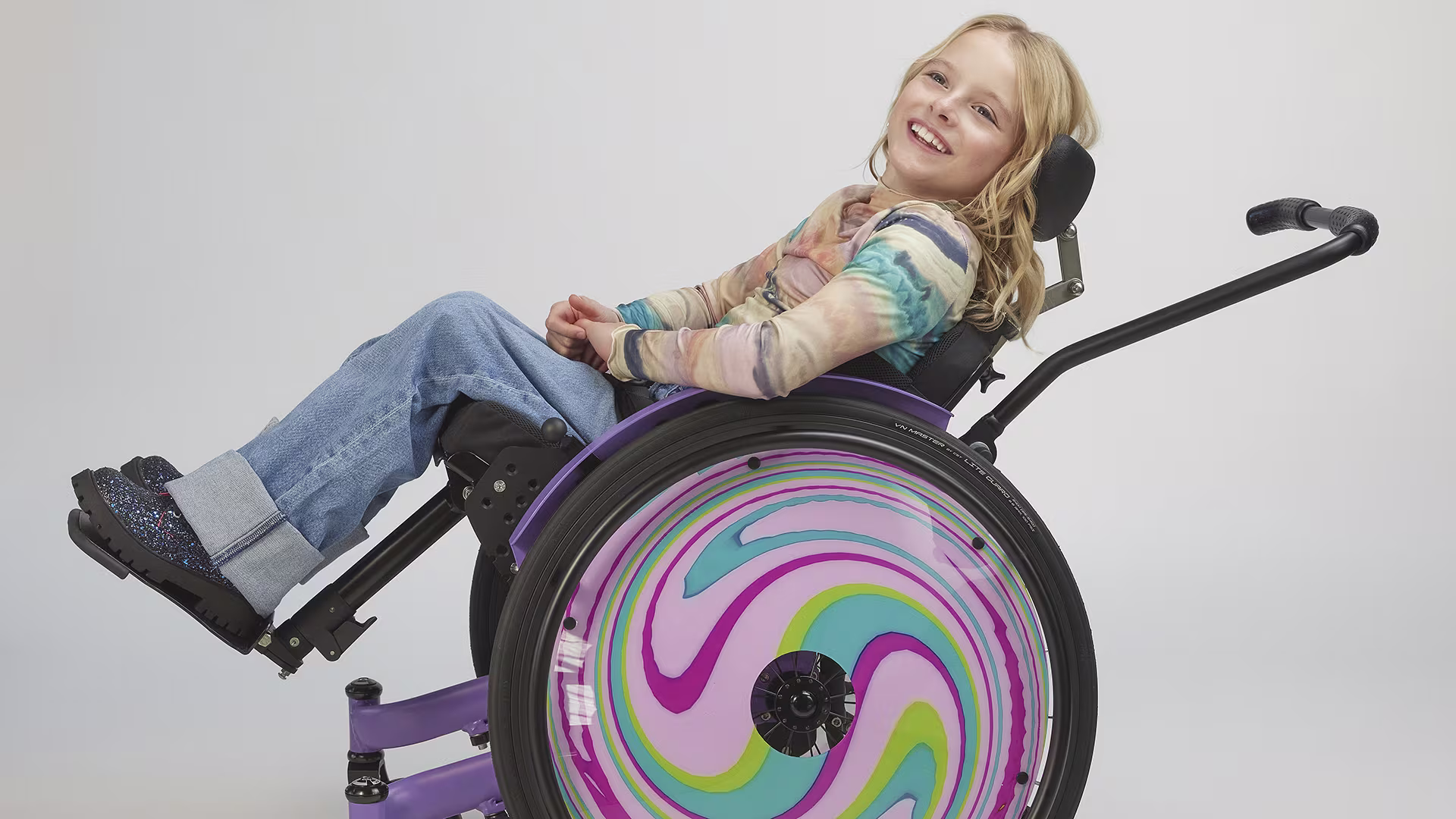 A blonde girl sits smiling in the Ottobock Kidevo children's wheelchair while the seat is tilted backwards