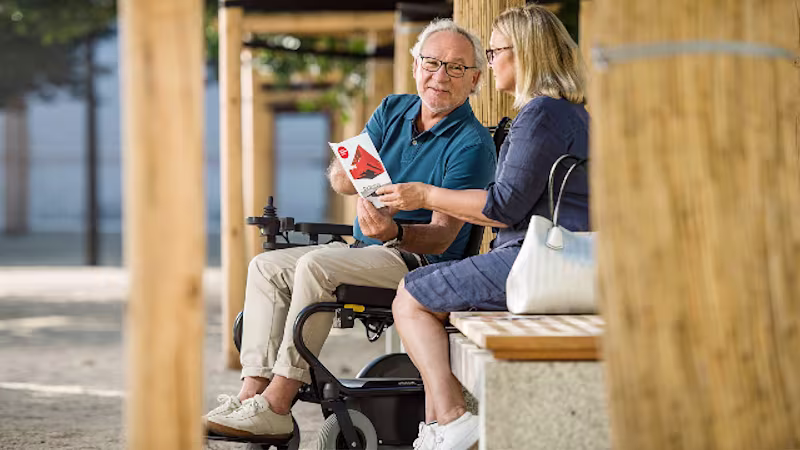 A man in a wheelchair sits next to a woman on a bench outside and shows her something in a brochure.