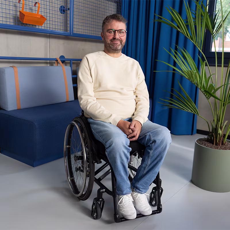 Eine männlich gelesene Person mit schwarz-grau melierten Haar und Dreitagebart sitzt in einem Avantgarde Rollstuhl von Ottobock. Im Hintergrund erkennt man eine blaue Kommode, ein blaues Sofa, blaue Vorhänge, einen roten Sessel und eine große Zimmerpflanze.