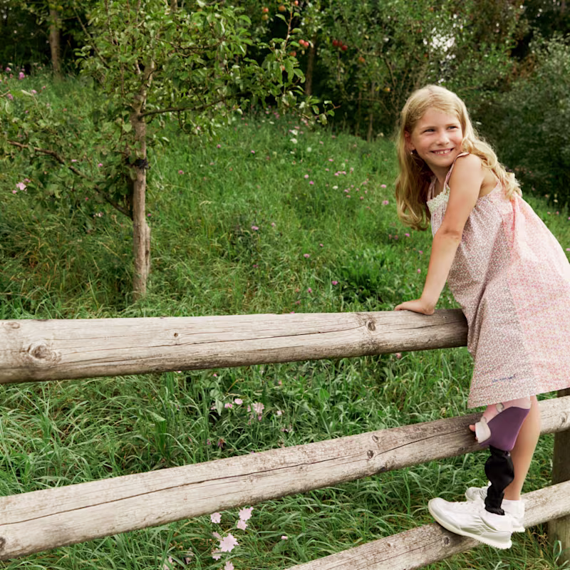 Auf dem Bild sieht man Nora, die auf einen Holzzaun klettert und die Prothesen Maverick junior & movido trägt.