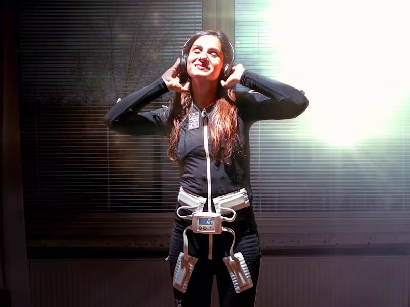 Person wearing Ottobock's Exopulse Mollii Suit and holding a cardboard virtual reality headset up to their face indoors.