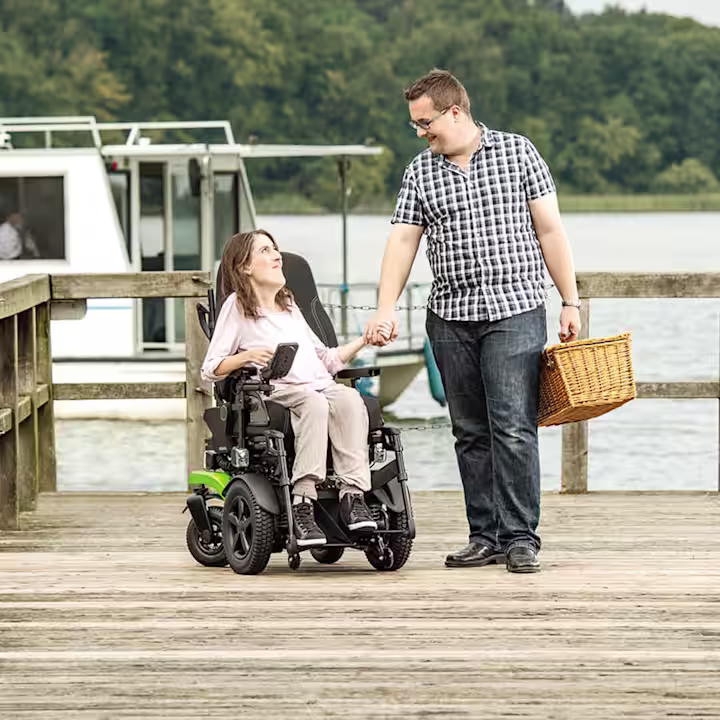 Eine weibliche Person sitzt im Juvo Elektrorollstuhl von Ottobock. Sie schaut ihren Mann liebevoll an und hält ihn an der Hand. Im Hintergrund ist ein Boot zu erkennen.