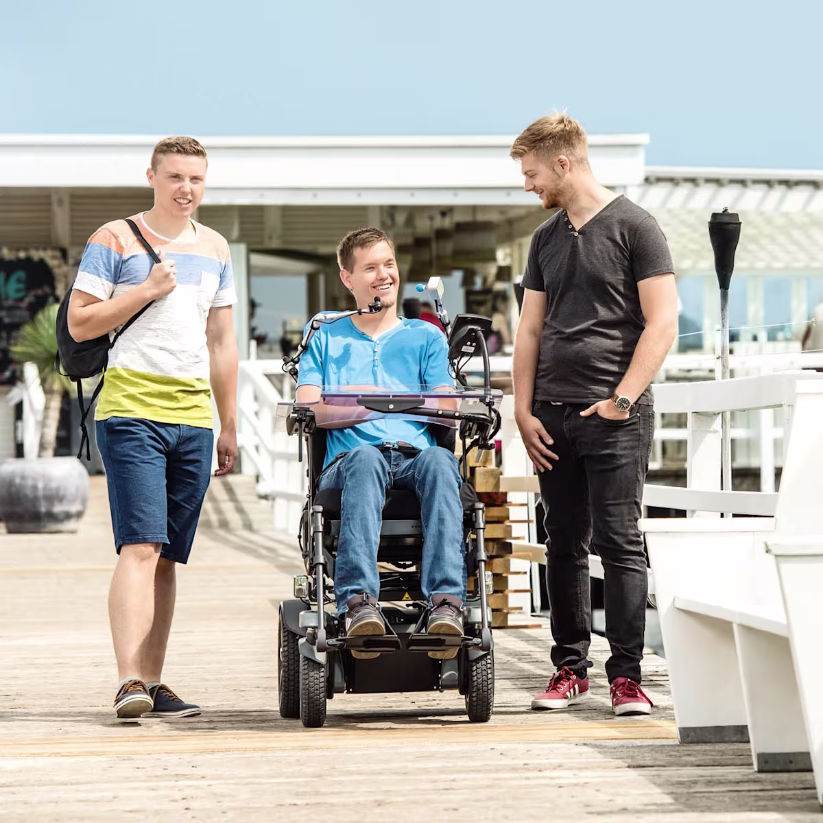 Ein junger Mann sitzt im Juvo Elektrorollstuhl. Links und rechts neben ihm stehen zwei Freunde von ihm. Sie befinden sich an einer Strandpromenade.
