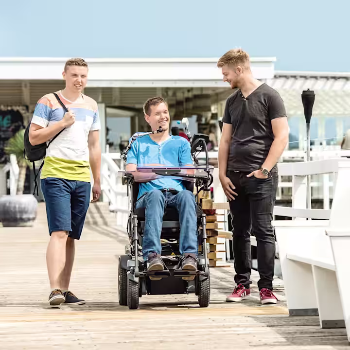 Ein junger Mann sitzt im Juvo Elektrorollstuhl. Links und rechts neben ihm stehen zwei Freunde von ihm. Sie befinden sich an einer Strandpromenade.