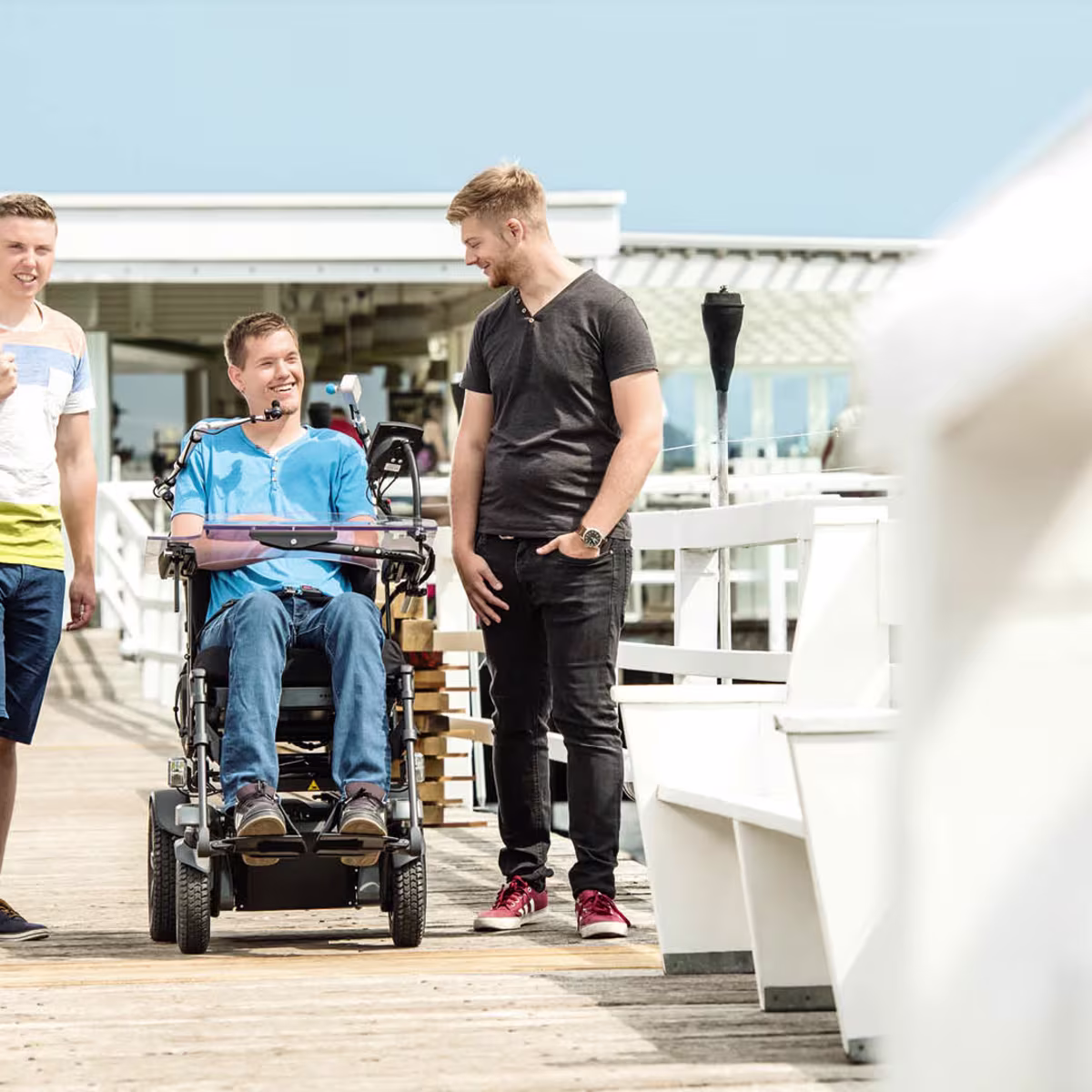 A young man is sitting in a Juvo power wheelchair. Two of his friends stand to the left and right of him. They are on a beach promenade.