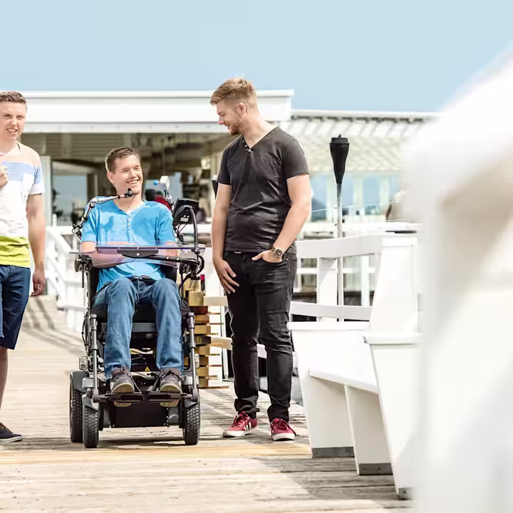 A young man is sitting in a Juvo power wheelchair. Two of his friends stand to the left and right of him. They are on a beach promenade.