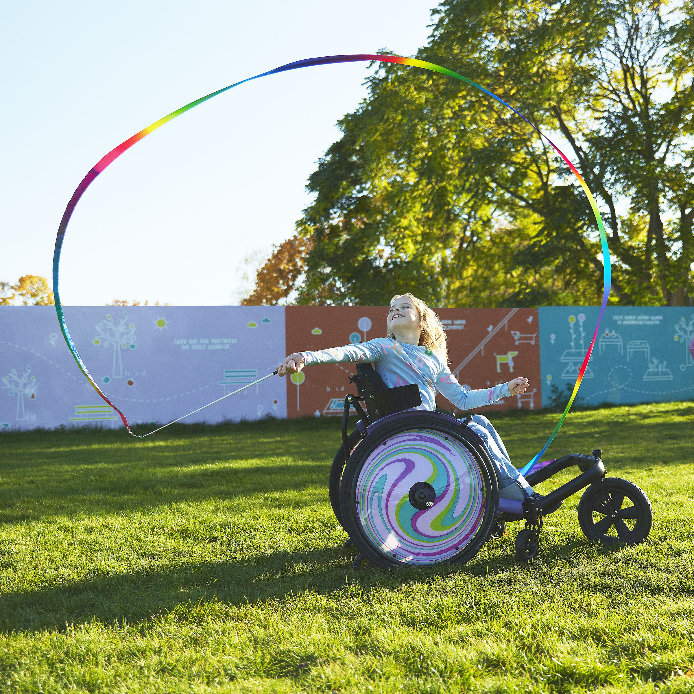 Genç bir kız, Nomine, kidevo tekerlekli sandalyesinin üzerinde renkli bir kurdeleyi döndürüyor. Jant kapakları mor ve mavi girdap desenine sahip.