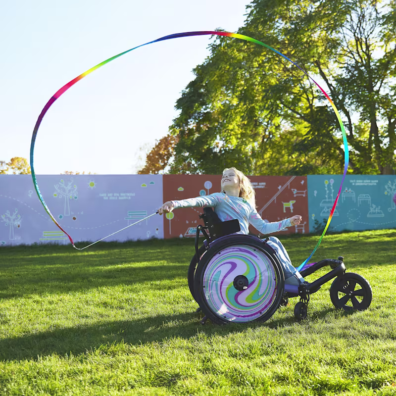 Genç bir kız, Nomine, kidevo tekerlekli sandalyesinin üzerinde renkli bir kurdeleyi döndürüyor. Jant kapakları mor ve mavi girdap desenine sahip.