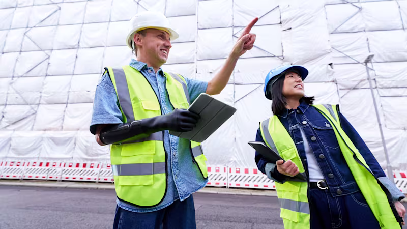 A prosthesis user holds a tablet with their prosthetic hand while standing with a colleague at a construction site.