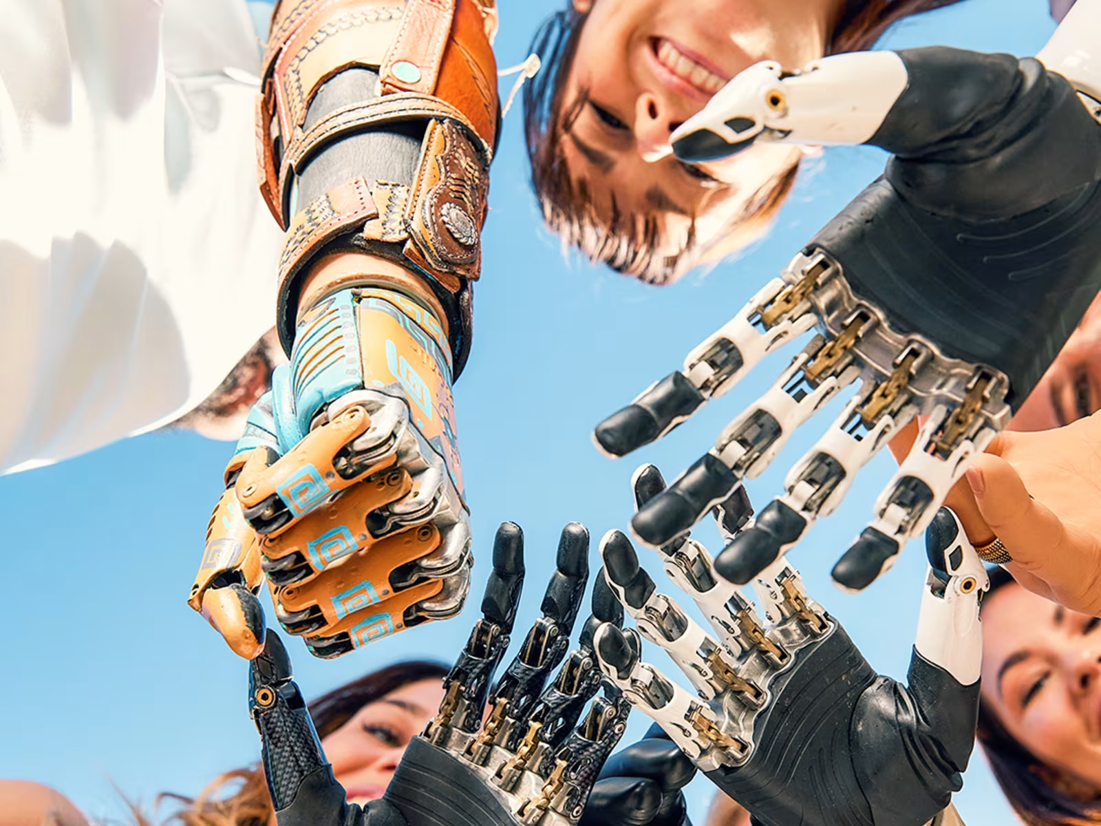 A group of smiling people standing in a circle, reaching their prosthetic hands toward the center against a bright blue sky, symbolizing community, technology, and empowerment.