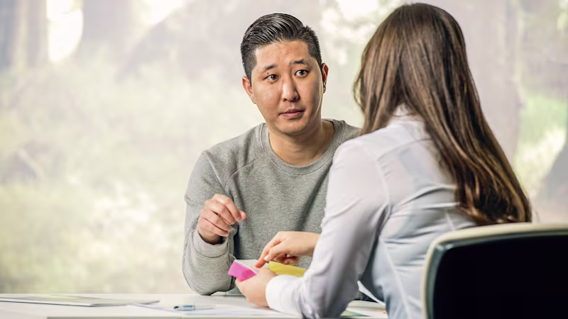 Male person sits at a table opposite a female person and listens to her while she points to a document