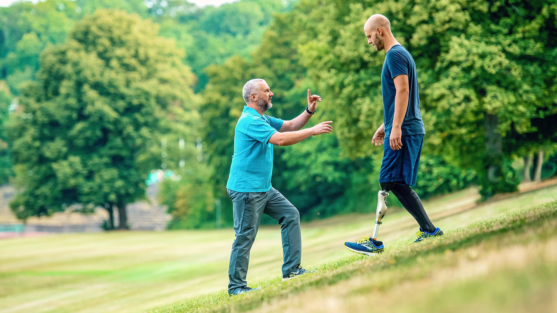 Amputee with Ottobock Kenevo microprocessor knee walking down a steep meadow while getting advice from prosthetist standing in front of him