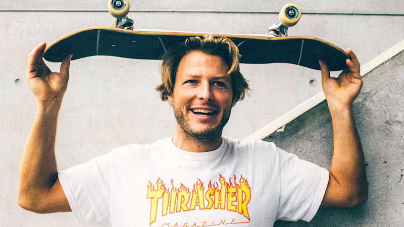A man smiles and holds a skateboard between his hands above his head.