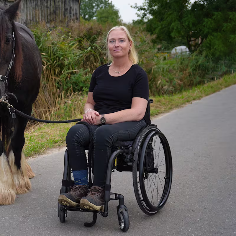 Eine weiblich gelesene Person mit blonden Haaren sitzt in einem Zenit Rollstuhl von Ottobock. Links neben ihr steht ein braunes Pferd.