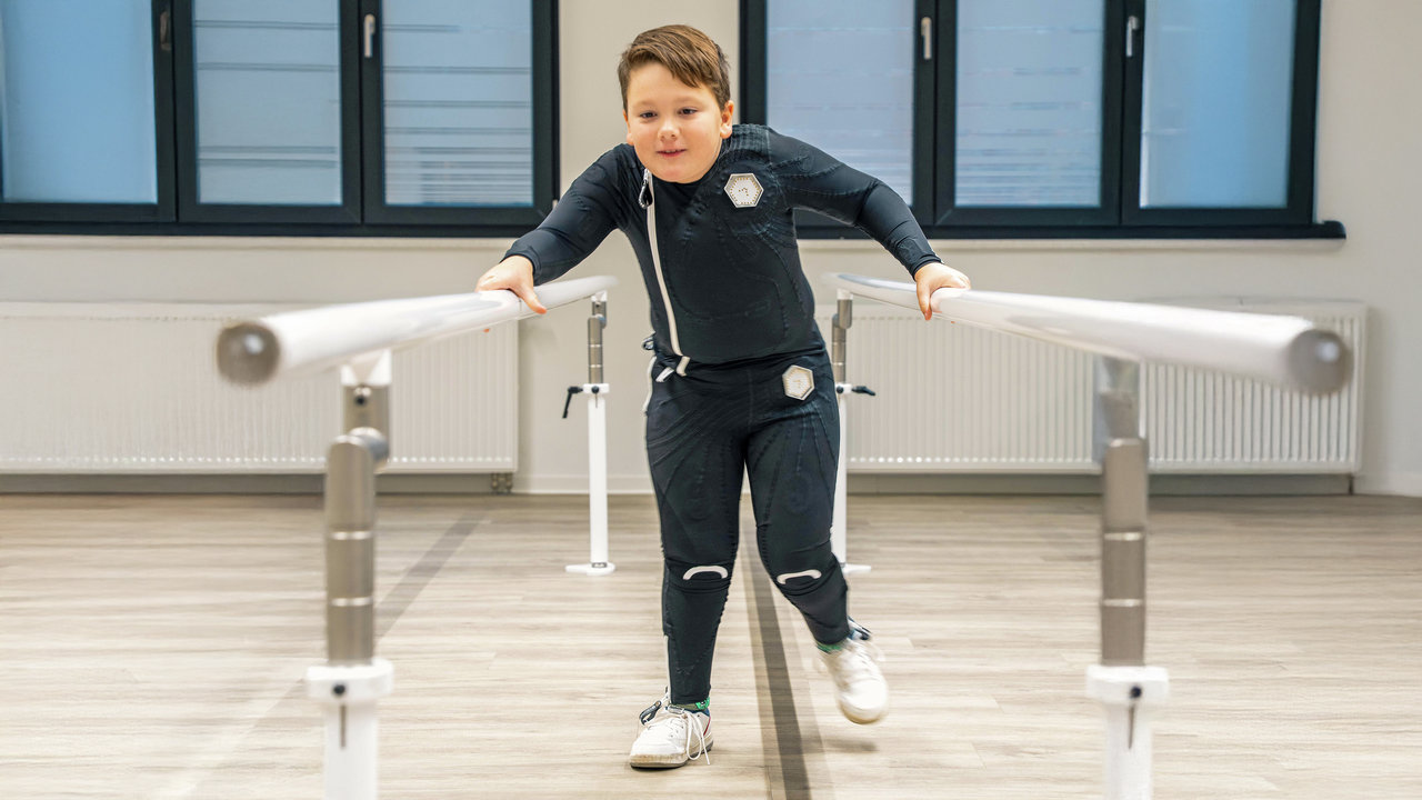 Ein Junge hat den exopulse suit von Ottobock an und traniert in einer Praxis sein Gangbild. 