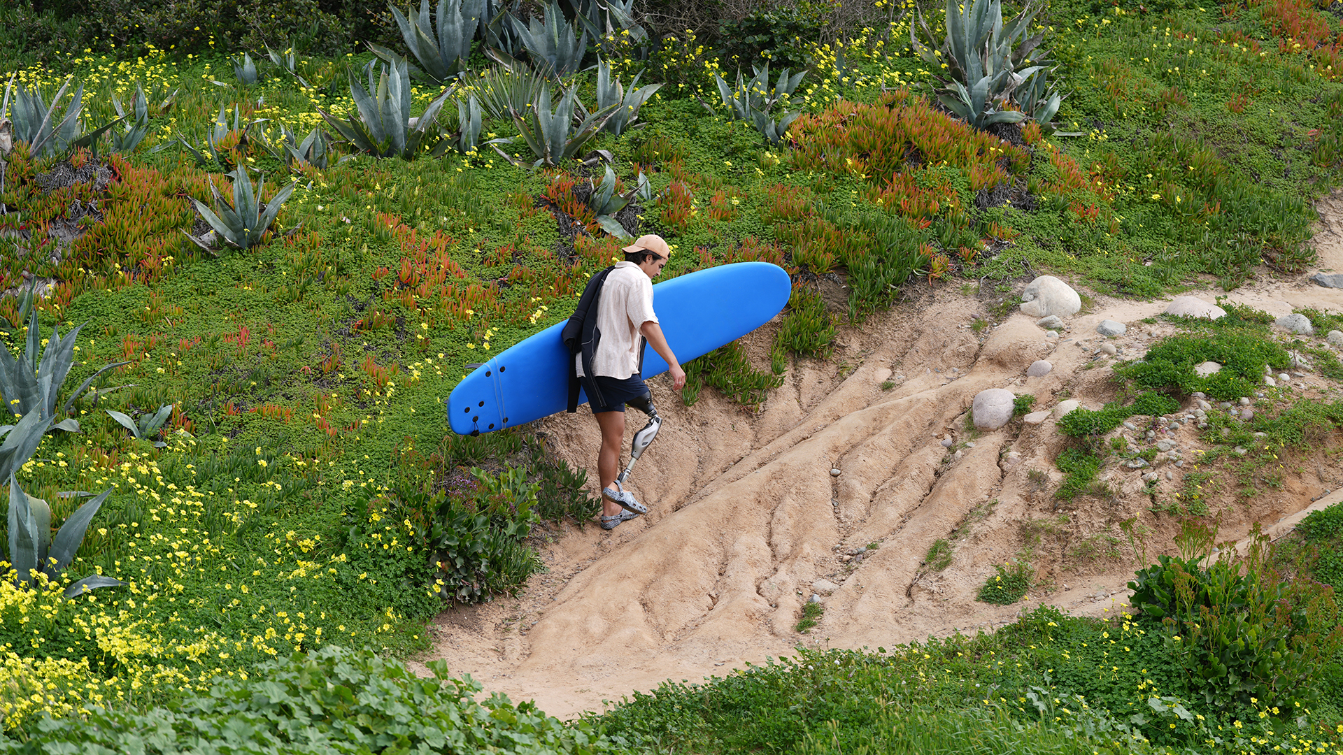 Genium X4 prosthetic knee user, Caleb, carrying his surfboard uphill along a beach