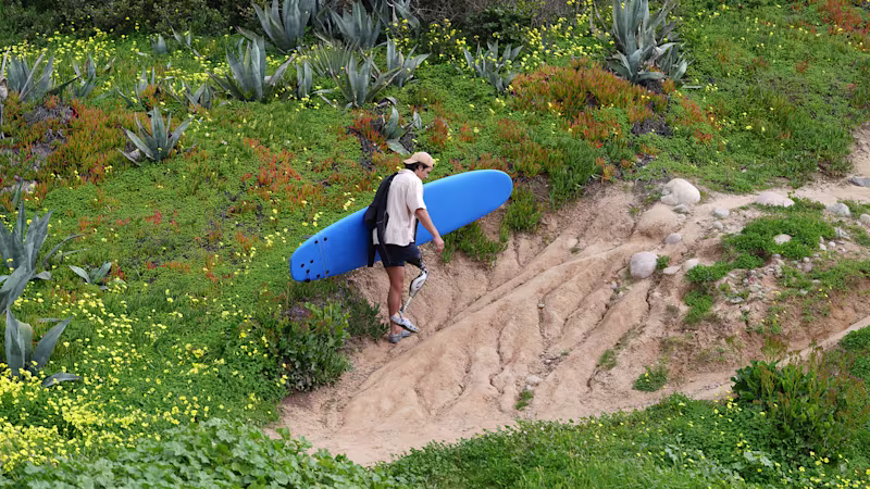 Genium X4 prosthetic knee user, Caleb, carrying his surfboard uphill along a beach