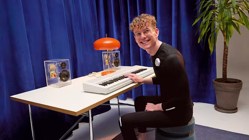 Person wearing an Ottobock exopulse suit smilingly playing Keyboard at a desk in front of a blue curtain