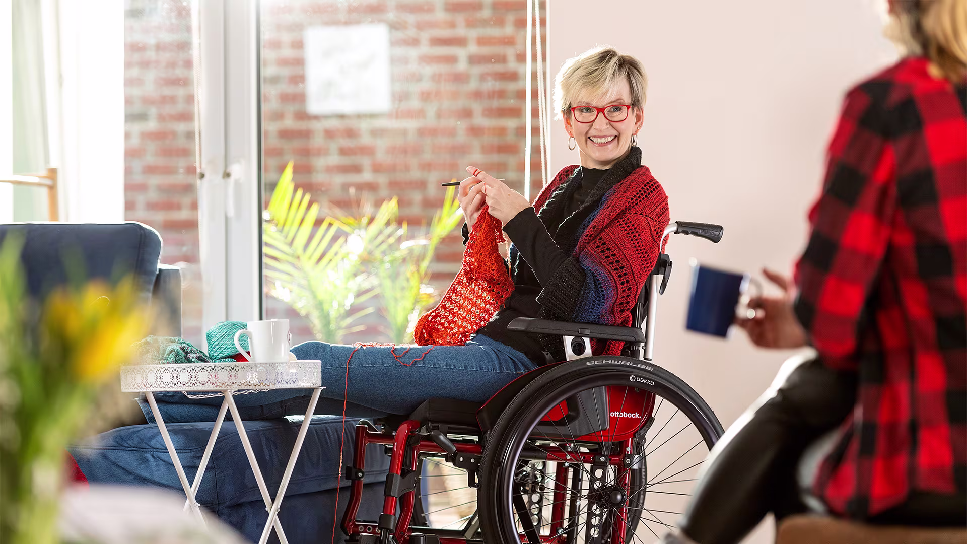 Woman in a Ottobock Motus wheelchair crocheting and looks smiling at person next to her