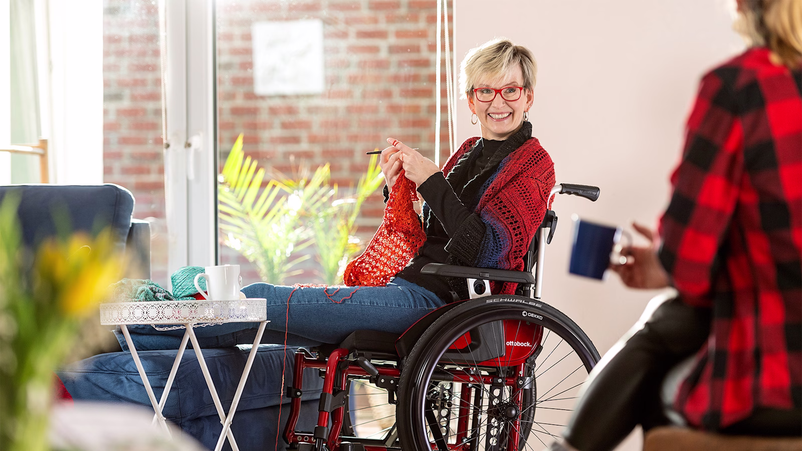 Woman in a Ottobock Motus wheelchair crocheting and looks smiling at person next to her