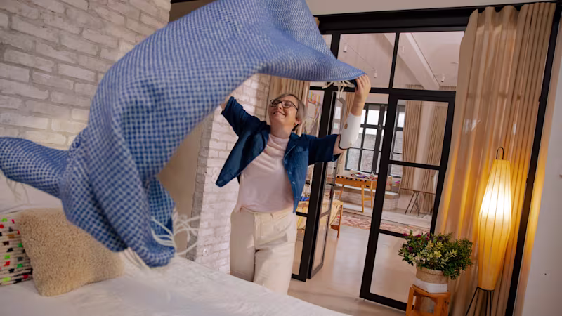 Eine Frau steht vor einem Bett und schüttelt freudig eine Tagesdecke aus, die sie auf das Bett legen möchte. Sie trägt eine Armprothese, die Speedhand.