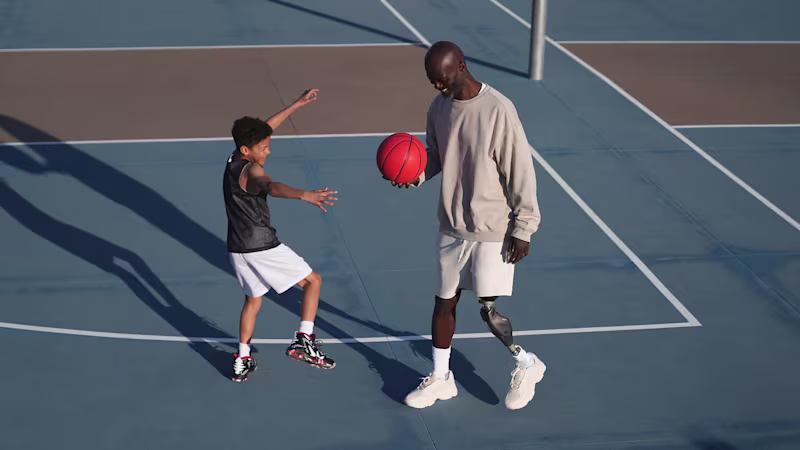 Ein Prothesenträger mit dem mechatronischen Prothesenkniegelenk Genium X4 von Ottobock spielt mit einem kleinen Jungen Basketball auf einem Basketballplatz.