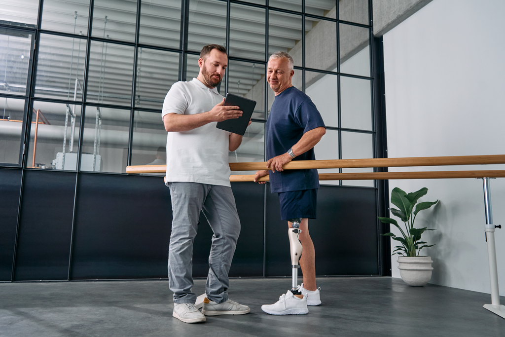 An orthopaedic technician explains Kenevo knee joint settings on a tablet while the user stands in parallel bars.