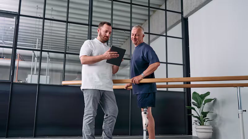 An orthopaedic technician explains Kenevo knee joint settings on a tablet while the user stands in parallel bars.