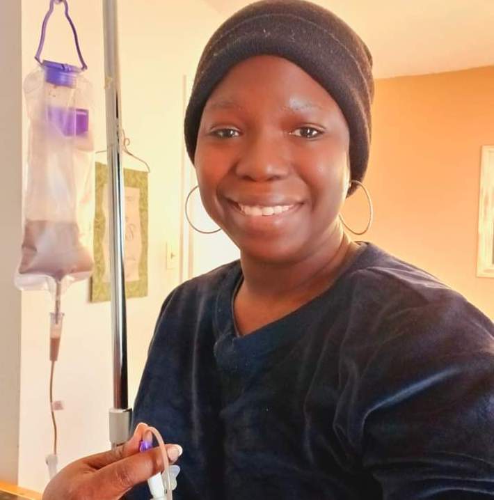 Photo of a women smiling wearing a beanie inside with a tube feeding bag hanging from a metal pole.