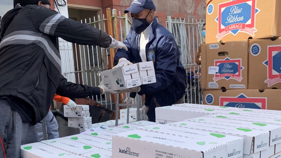 NYC Mayor, Eric Adams, wearing a mask, gloves and baseball hat, hands boxes of Kate Farms to a volunteer at the Campaign Against Hunger. He is standing amongst several stacks of boxes as other volunteers work in the background.
