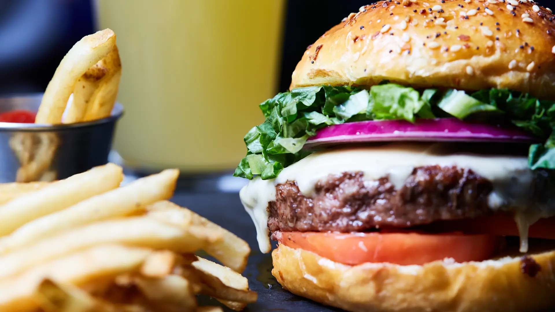 A close-up of a juicy cheeseburger with lettuce, tomato, onion, and melted cheese in a seeded bun, accompanied by crispy fries and a drink in the background.