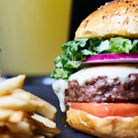 A close-up of a juicy cheeseburger with lettuce, tomato, onion, and melted cheese in a seeded bun, accompanied by crispy fries and a drink in the background.