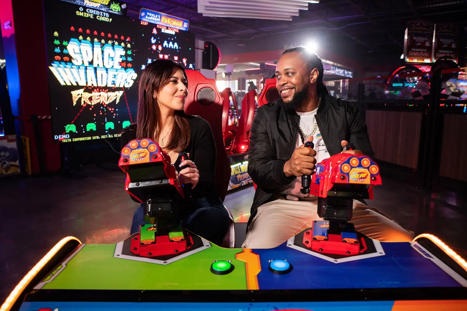 A woman and a man are sitting side by side at an arcade, playing a game together. Both are smiling and focused on the game. In the background, there are bright neon lights and a large "Space Invaders" game.