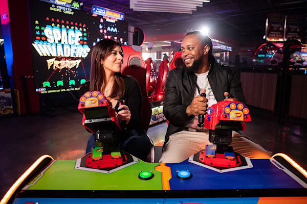 A woman and a man are sitting side by side at an arcade, playing a game together. Both are smiling and focused on the game. In the background, there are bright neon lights and a large "Space Invaders" game.