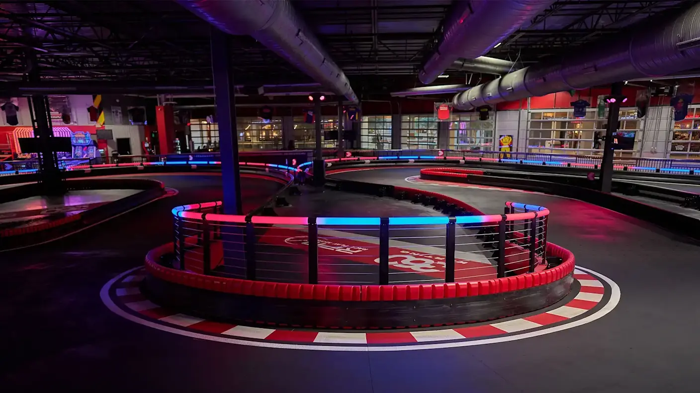Indoor go-kart racetrack in Syracuse with neon red and blue lighting. The track has several curves and turns, surrounded by protective barriers. The ceiling features exposed ducts and pipes, and there are large windows in the background showcasing different areas.