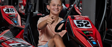 A young boy wearing a light pink t-shirt and gray shorts is sitting in a red go-kart with the number 52. He is smiling and giving a thumbs-up gesture. The background shows more go-karts with numbers, indicating an indoor go-kart facility.