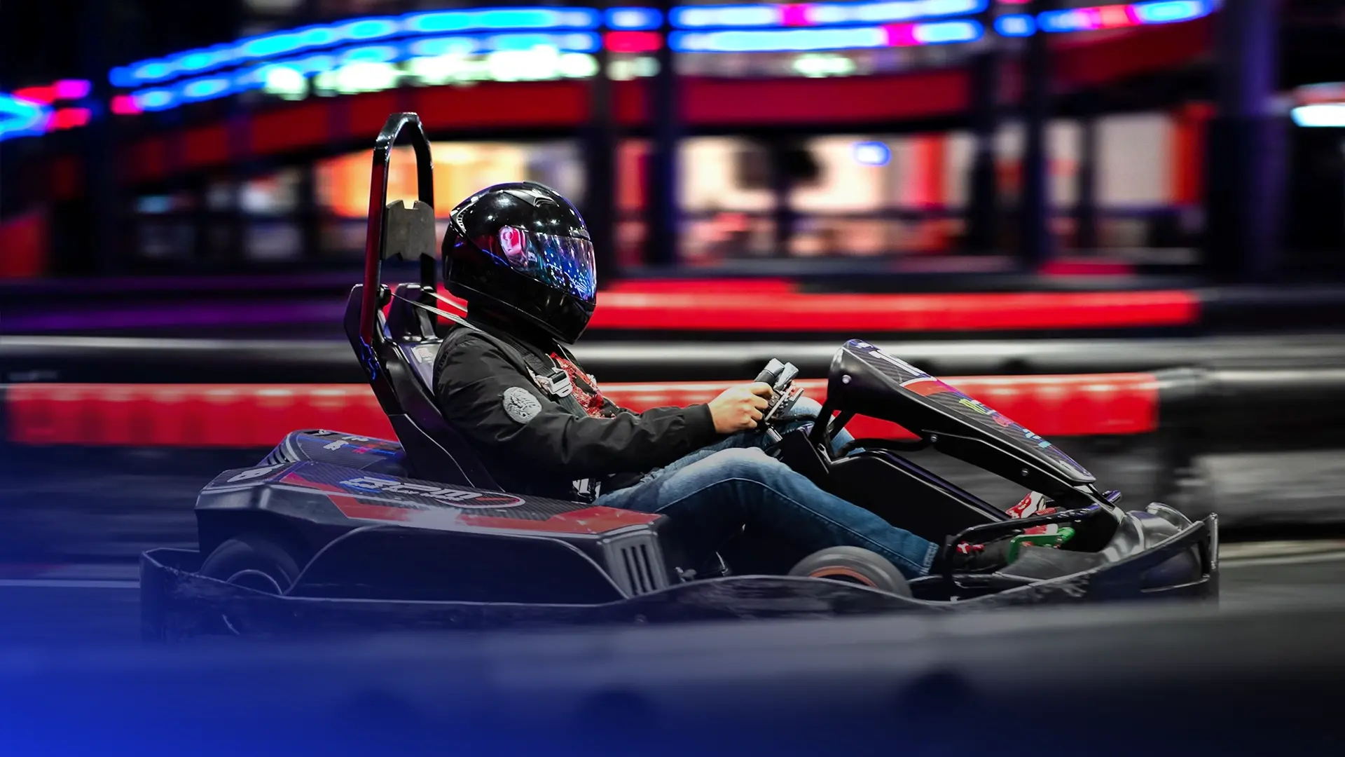 A person wearing a black helmet and jacket drives a go-kart on an indoor track. The background is blurred, highlighting the red and black barriers and vibrant neon lights, creating a sense of high speed.