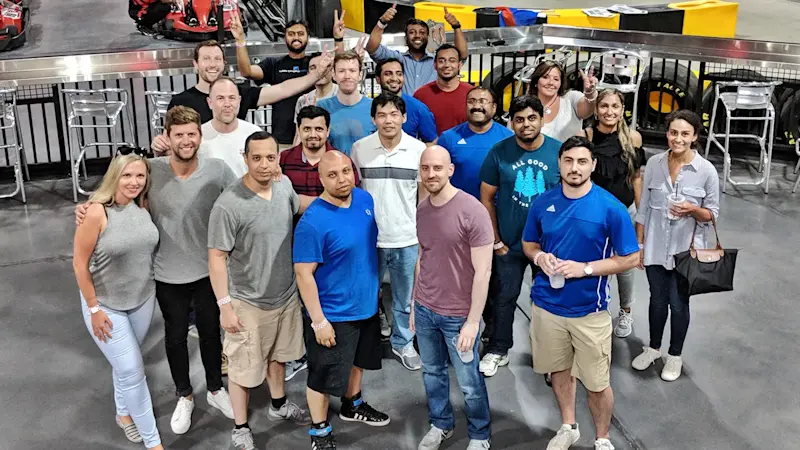 A diverse group of people, casually dressed, stands together in an indoor space next to a metal railing. Some people are holding drinks and smiling at the camera. In the background, go-karts and a racing track are visible.