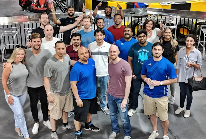 A diverse group of people, casually dressed, stands together in an indoor space next to a metal railing. Some people are holding drinks and smiling at the camera. In the background, go-karts and a racing track are visible.