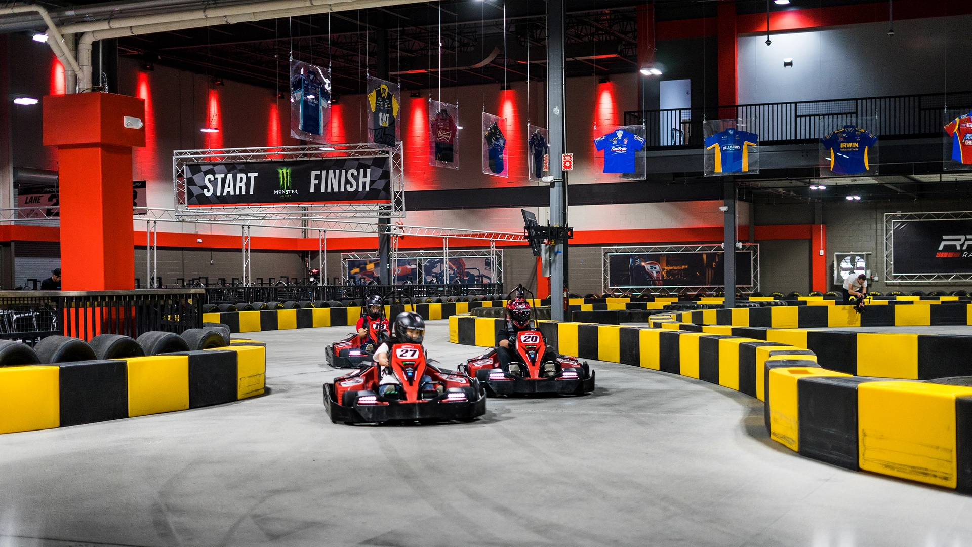 Indoor go-kart racing track called RPM with three go-karts on the course. The track has black and yellow barriers, and there is a "Start" and "Finish" banner hanging overhead. The background features a viewing area with shirts displayed on the railing.