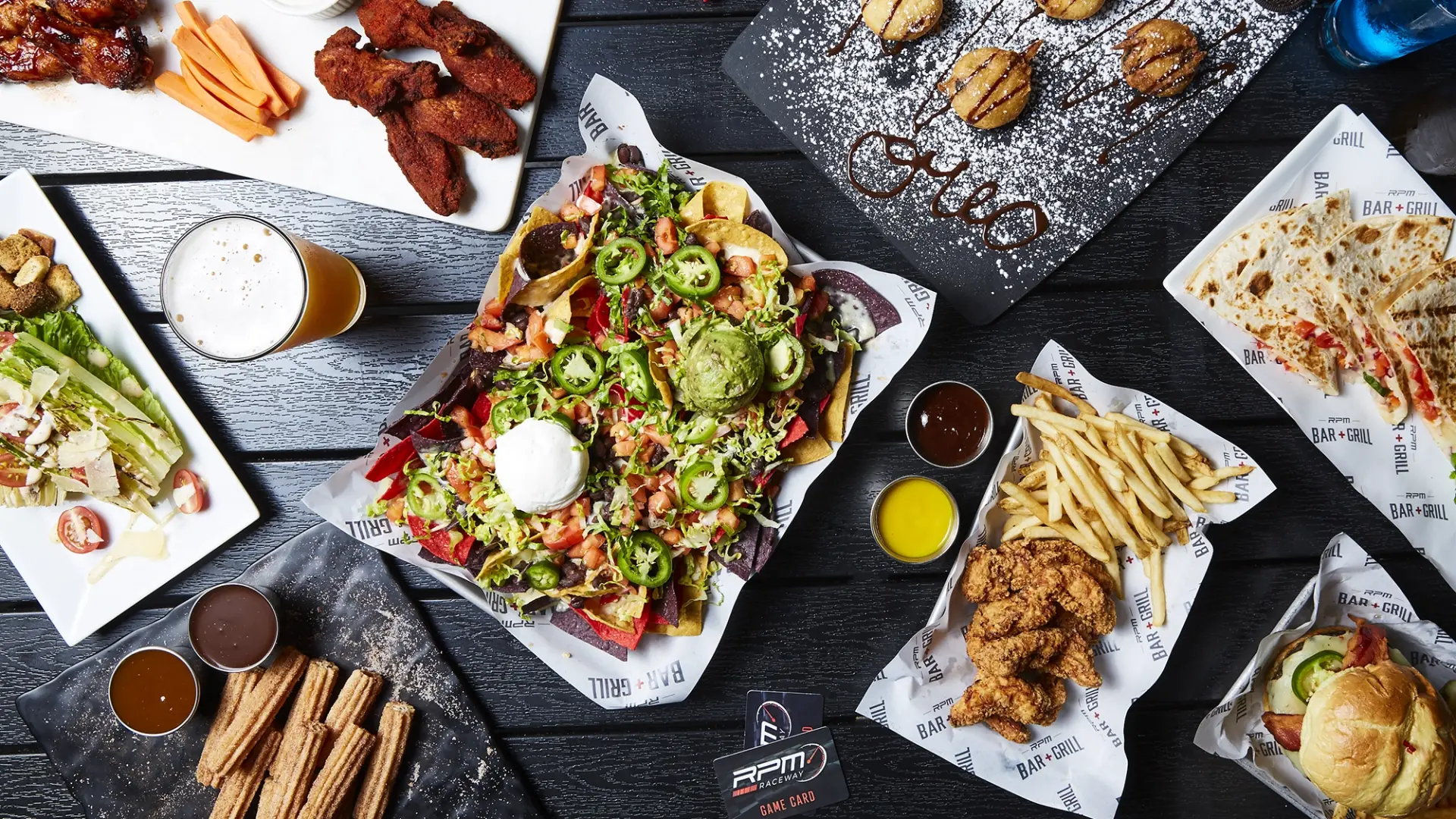 A top view of various dishes on a dark table, including a plate of nachos topped with jalapeños, guacamole, and sour cream, a quesadilla, seasoned french fries, chicken wings, and a glass of beer. Smaller plates hold desserts, fried items, and dipping sauces.