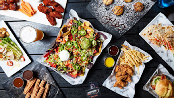 A top view of various dishes on a dark table, including a plate of nachos topped with jalapeños, guacamole, and sour cream, a quesadilla, seasoned french fries, chicken wings, and a glass of beer. Smaller plates hold desserts, fried items, and dipping sauces.
